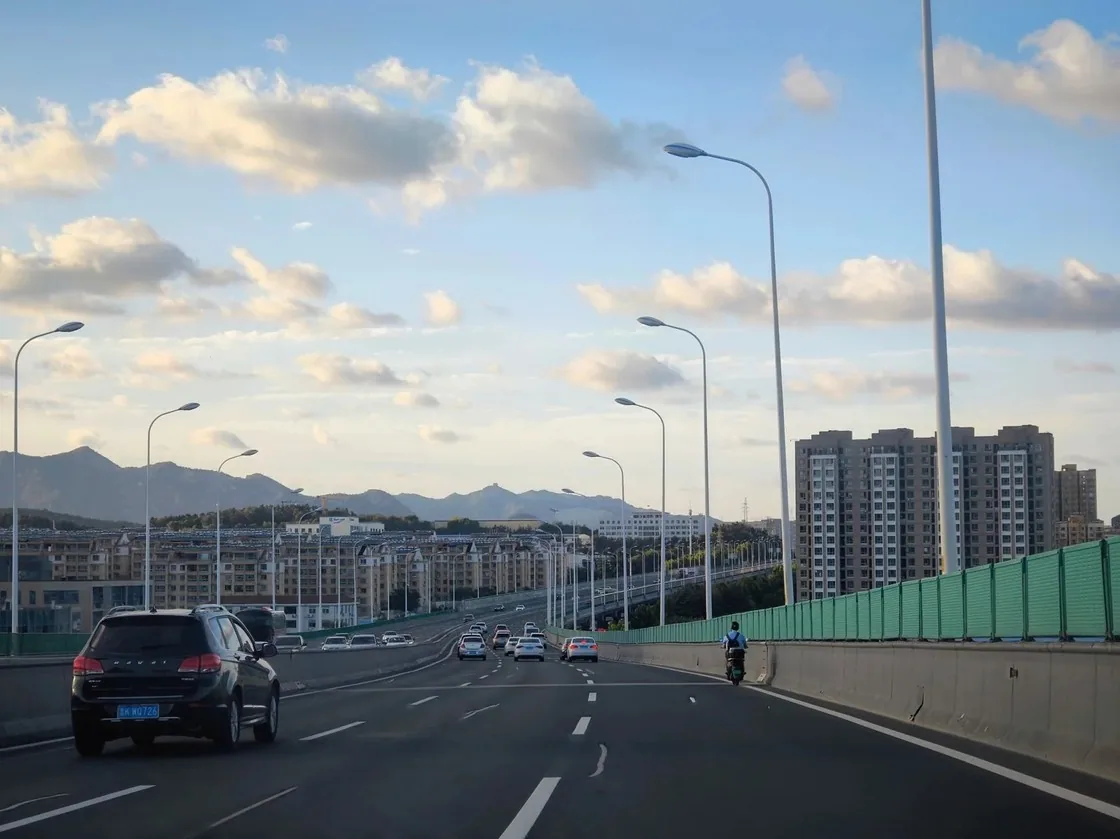 高架路上车流向前,威海市区的楼群与远山在蓝天白云下迎面铺开