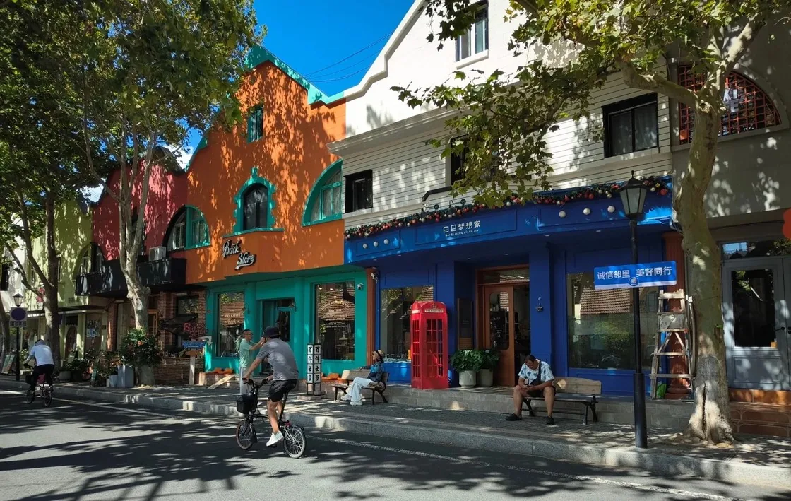 新威附路街景,橙色与蓝色的彩色小屋沿街排开,门口立着红色电话亭,梧桐树影洒满路面